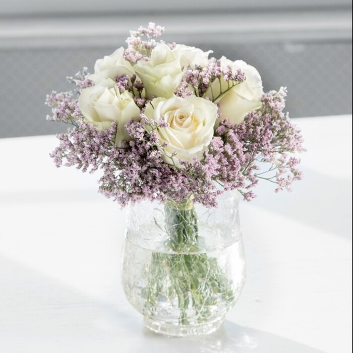 A beautifully balanced bouquet of white roses and lilac statice is the clear focal point of this image, captured in bright, diffused daylight that pours in from an unseen window. The arrangement is placed in a tall, transparent glass vase set on a pristine white surface, echoing the clean, modern style often seen in homes around Gerrards Cross and the nearby Denham Lane area. About twelve creamy white roses crown the design, each flower showing layers of velvety petals that curl gently outwards, so that no bloom is fully open yet none appear tight or closed. Their hue sits gracefully between soft ivory and warm cream, giving them a refined, almost luminous quality as the light brushes their contours and edges. Nestled among and around these roses, airy sprays of light lavender statice spread in every direction, their tiny clustered flowers creating a gauzy, textural surround. The statice lends the bouquet a more relaxed and slightly whimsical feel, preventing the classic roses from appearing too formal or severe, and the cool purple tones offer a quiet contrast to the warmth of the white blooms. Looking through the clear glass of the vase, you can see the gathered green stems rising straight and fresh from the base, neatly trimmed and submerged in crystal-clear water that sparkles softly. The background is softly blurred, ensuring the viewer's attention remains on the flowers while still hinting at a calm interior, perhaps a dining table ready for guests or a console in a peaceful hallway. This composition suggests gentle emotion-ideal for a sympathy gift, a tasteful office arrangement for a practice near East Common, or a simple expression of affection-where the message is delivered with understated elegance and sincere, enduring beauty.