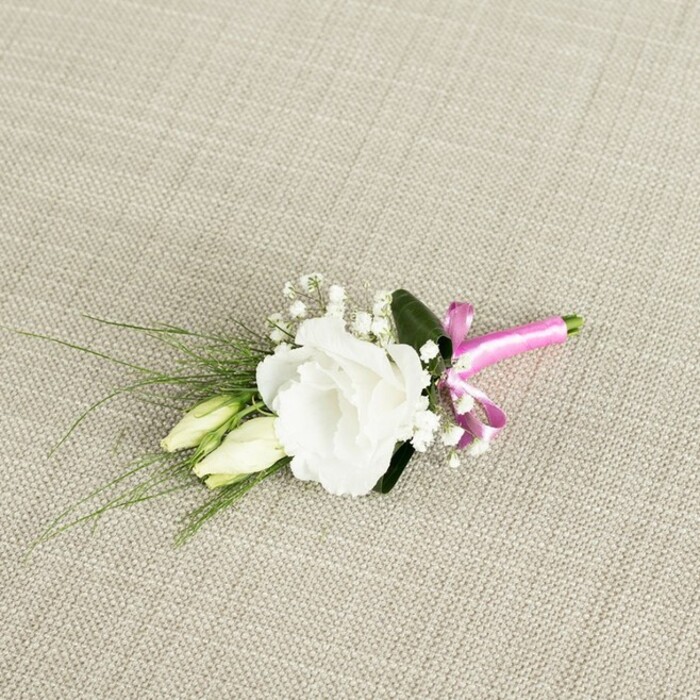 A small yet exquisitely composed floral boutonnière is captured from a slightly elevated angle, resting diagonally across a light beige, finely woven fabric that evokes the texture of natural linen or softly worn burlap. At the centre of the design is a fully opened white eustoma, or lisianthus, its many ruffled petals unfolding in soft layers, creating a rounded, almost rose-like form. The petals transition from a crisp, luminous white at the edges to gentle shadows nearer the throat of the bloom, giving it a sense of depth and delicacy that would suit a refined wedding in Gerrards Cross perfectly. Just to the left of this central flower are two unopened lisianthus buds, slender and elongated, their creamy-white forms brushed with pale green, hinting at flowers that will open later in the day. Interspersed among the main bloom and buds are airy stems of white baby's breath, their tiny, star-shaped blossoms offering a light, cloud-like texture that fills the gaps without overwhelming the composition. Radiating from behind the flowers, slim, grass-like blades of deep green foliage sweep toward the lower left corner, giving the boutonnière a graceful, organic line, while a broader, darker green leaf tucked behind the central flower adds contrast and visual weight. The stems are carefully bound at the base with a vivid fuchsia pink satin ribbon, tied into a petite, neat bow whose silky sheen catches the light and introduces a playful, modern accent to the classic palette. The neutral background and soft, even lighting ensure that every nuance of petal, leaf, and ribbon is clearly visible, making this piece ideal for a groom or prom guest dressing for a celebration anywhere from the leafy avenues near East Common to intimate gatherings across the wider Gerrards Cross area.