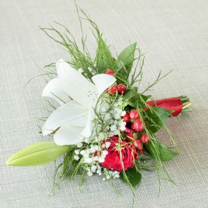Seen from above as if resting on a sunlit window ledge in Gerrards Cross, this refined boutonniere or miniature bouquet is a testament to the skill and passion of local florists. A crisply unfurled white lily takes centre stage, its large petals pristine and lightly speckled with warm earthy reds near the heart, summoning visions of lilies in bloom along West Common's borders. Just below, a rich, velvet-red rose blooms fully in dramatic contrast, its petals densely layered in a traditional spiral that suggests enduring affection. Petite white baby's breath catches the light, weaving softness and brightness throughout and reminiscent of wildflowers dotting Bulstrode Park. Vivid clusters of round, red berries glimmer, lending a festive spirit to the piece. A single, gentle green lily bud stretches to one side, promising new life amid lush abundance. The bouquet's greenery-wispy asparagus fern, elegant dark leaves-enfolds the flowers, adding a woodland feel and texture. Everything is expertly tied with a bold, lustrous red satin ribbon, perfectly echoing the crimson of rose and berry. Thoughtfully arranged for special occasions in Gerrards Cross, such as weddings or anniversaries, this floral accessory radiates sophistication, heartfelt celebration, and the artistry of bespoke local floral design.
