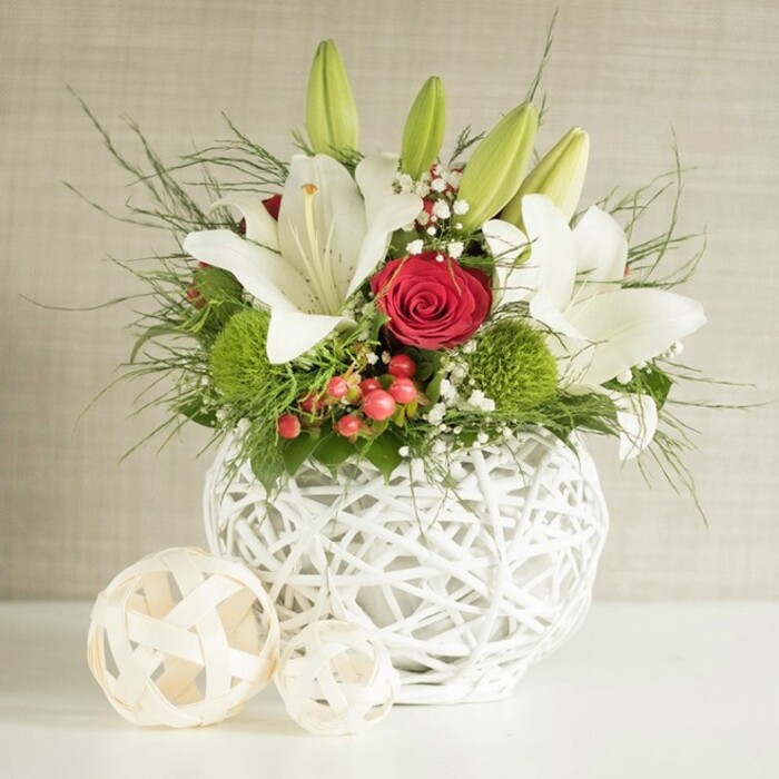 This image captures a sophisticated floral centerpiece that could easily grace a wedding table at a boutique hotel or private home in Gerrards Cross. The arrangement is styled in a round white vase made from interwoven strands that create an open, basket-like texture, giving a sense of handcrafted artistry and lightness. From this contemporary container rises a full, rounded bouquet where pristine white lilies are the stars. Some lilies are wide open, with broad, velvety petals revealing golden stamens and gentle speckles near the throat, while several buds remain closed, their elongated green forms arching outward and hinting at days of continuing bloom and fragrance. Among the lilies, deep red roses provide a rich counterpoint, the most prominent one positioned near the centre with its velvety petals tightly folded into a spiralled heart, evoking romance and timeless elegance. Between these main blooms, bright green pom-pom flowers-likely green chrysanthemums or dianthus-add a unique spherical form and a fresh shock of colour, reminiscent of the lush greens in nearby Bulstrode Park. Fine clusters of white baby's breath are scattered throughout, giving the arrangement an airy, cloud-like softness. Small sprays of red berries tuck into the foliage, suggesting celebration and adding a seasonal touch without overwhelming the design. A variety of greenery, from slender, feathery fronds to darker, broader leaves, spills naturally around the flowers, softening the edges and providing depth. On the white surface beside the vase, two decorative woven spheres in a pale wood tone reinforce the natural theme and mirror the open-lattice pattern of the vase. The backdrop is a subtly textured wall in light beige or grey, quiet and understated, allowing every petal, leaf, and berry to stand out clearly, as if inviting guests in Gerrards Cross to pause and admire the craftsmanship.