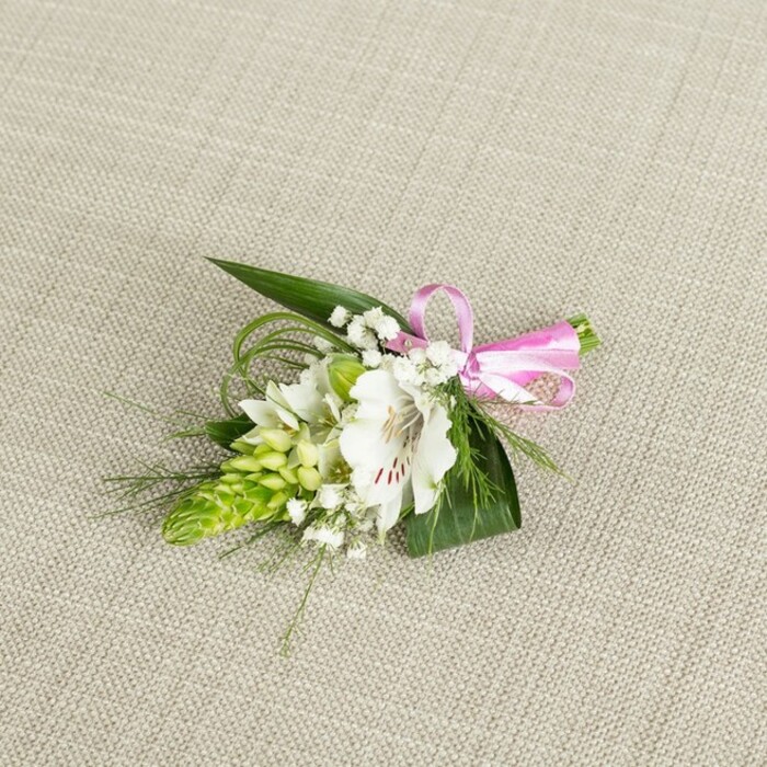 An elegant, small-scale floral boutonniere is displayed on a softly textured, light beige surface that looks like fine linen draped across a table in a Gerrards Cross townhouse. At the centre of the composition sits a single white alstroemeria, or Peruvian lily, its petals smooth and slightly curved, opening wide to reveal a throat flecked with delicate reddish-brown speckles that run like fine brush lines from base toward the mid-petal. The bloom appears fresh and just fully open, its stamens subtly visible, giving a feeling of vitality and poised beauty. Nestled alongside is a vivid lime-green bud cluster, reminiscent of an Ornithogalum or ginger-like spike, with numerous tightly packed buds stacked in a tidy, conical formation, contributing both structure and a lively green contrast. Threaded throughout the piece are ethereal sprigs of white baby's breath, each minuscule bloom poised on slender stems, creating a gauzy, cloud-like frame around the central lily. The flowers are cradled by lush, dark green, pointed leaves that fan outward and provide a strong visual base, while fine, grass-like blades loop and arc gracefully, hinting at natural movement, as though caught in a gentle Chiltern breeze from nearby Bulstrode Park. At the upper right, a neat bow of glossy, bright pink satin ribbon secures the stems, its soft sheen and cheerful colour injecting a playful, romantic note without overwhelming the delicate white and green palette. The lighting is even and calm, with no harsh contrasts, allowing every texture-from the weave of the fabric background to the smoothness of the petals-to be clearly perceived. This thoughtful boutonniere feels ideal for a wedding, prom, or anniversary celebration in Gerrards Cross, embodying refined craftsmanship and the intimate joy of a special day shared with loved ones.