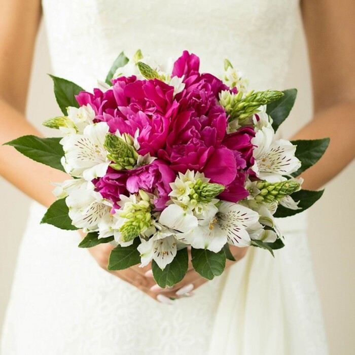 In this intimate wedding photograph, the camera lingers on a hand-tied bouquet held close to the midsection of a bride dressed in a white gown, the delicate texture of lace and smooth fabric forming a luminous backdrop. At first glance, the bouquet reads as a strong, romantic burst of colour: large fuchsia and magenta peonies cluster at the centre, their many ruffled petals layered so tightly that each bloom looks like a small, luxurious pom-pom of silk. Between these generous blossoms, white alstroemeria unfurl with quiet elegance, each petal slim and slightly curved, marked inside with fine maroon or deep burgundy streaks that feel almost like calligraphy. Rising up through the composition are narrow, spiky green buds and textural florets, adding lively vertical accents that keep the rounded shape from appearing too static. Around the outside, rich, dark-green leaves form a natural frame, their surfaces glossy and smooth, echoing the freshness of a garden after rain in nearby Bulstrode Park. The bride's hands are visible just below the blooms, fingers wrapped gently around the stems, nails painted a soft pink that harmonises with the deeper magenta tones above. The background fades into a creamy blur, likely the interior of a local church or a reception venue around Gerrards Cross, so the eye stays fixed on the interplay of colour and texture in the flowers. The bouquet feels dense yet airy, full of movement and life, suggesting the faint sweetness of peonies and the green, slightly herbal scent of freshly cut foliage. This image encapsulates the spirit of a modern Gerrards Cross bride - confident, romantic, and drawn to bold, joyful colour balanced with classic white - making it an inspiring reference for anyone planning a spring or early summer wedding in the area.