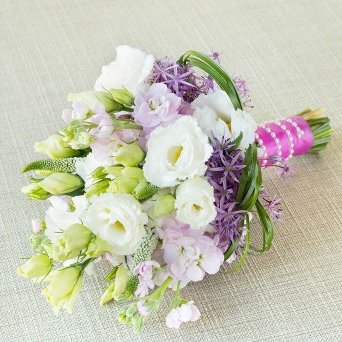 Resting diagonally upon a muted, textured beige backdrop reminiscent of fine linens in Gerrards Cross's stately homes, this bouquet exudes an air of natural grace. The arrangement leans heavily on snowy white Lisianthus, their ruffled petals unfurling with an inviting softness, each flower crowned by a gentle pale yellow center. Accentuating their purity, sprays of tender pale green buds and petite, elongated Lisianthus combine with soft clouds of lavender-pink stock, whose delicate fragrance adds romance and layer. What truly sets this bouquet apart are the dramatic deep purple allium blooms, their radiant, star-like florets jutting outward to create sculptural contrast-echoing the symmetry of well-manicured gardens along West Common. Slender, vivid green bear grass loops fluidly through the assemblage, imparting a sense of movement akin to the breezes through Gerrards Cross's surrounding meadows. Dense, textured fillers and hints of youthful green growth further amplify the feeling of renewal. Binding the stems is a luxurious fuchsia satin ribbon, crisply entwined with lustrous white pearls, providing both a springtime flourish and time-honoured sophistication. The soft, ambient lighting enhances the pastel palette and invites contemplation-this is a bouquet for capturing memories, ideal for elegant weddings, anniversaries, or thoughtfully curated local gatherings in Gerrards Cross. Its blend of refined whites, vibrant purples, and lively greens offers a fresh interpretation of classic romance, perfect for any event infused with a sense of community and artistry.