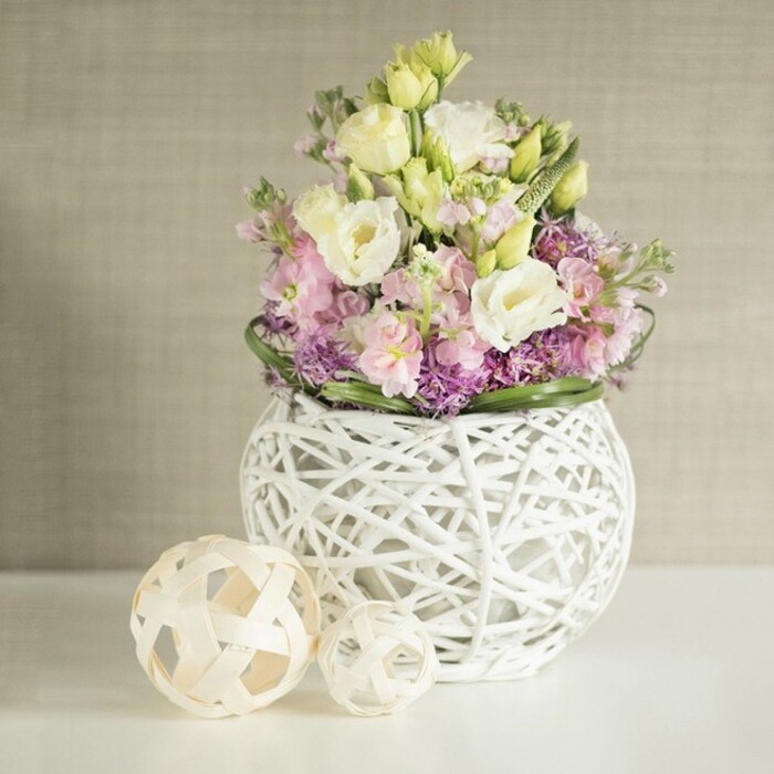 A still life imbued with understated elegance, this artfully arranged bouquet captures the refined yet natural spirit of Gerrards Cross living. The main feature is a lavish collection of creamy eustoma, with their softly ruffled petals layered alongside blush pink and lilac matthiola, each stem rising to add grace and height, reminiscent of early blooms along the Misbourne during spring. Small clusters of vibrant purple fillers nestle among the central blossoms, lending depth and texture reminiscent of local wildflowers. Accents of fresh green foliage and pale buds peek through, completing the picture of abundance. All are gathered into a distinctive, spherical white vase crafted from openwork painted rattan, its airy, woven structure exuding both modern style and rustic charm-echoing artisanal touches found in Gerrards Cross homes. To the left, two elegantly crafted spheres made from natural rattan, their forms echoing the vase's basket-weave motif, rest atop a crisp white tabletop that amplifies the light and colours of the scene. The background-a warm, softly textured greige wall covering-serves as a subtle yet refined canvas, highlighting the gentle palette of the arrangement. Perfect for wedding tables at local venues or as an inviting focal point for a Denham Lane residence, the bouquet effortlessly captures a mood of gentle celebration, bespoke craftsmanship, and the freshness of the local countryside, making it an inspiring reference for articles on seasonal decor, romantic centrepieces, or the artistry of Gerrards Cross floristry.