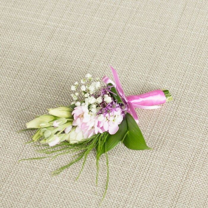 A meticulously designed boutonniere or petite corsage is positioned centrally on a pale, oatmeal-coloured linen surface, its fine weave and subtle texture echoing the understated elegance of many homes and venues around Gerrards Cross. The focal point of the piece is a small cluster of soft pink blooms, likely miniature stocks or similarly ruffled garden flowers, layered with countless petals that curl and fold in gentle waves. Their colour moves from a delicate, almost white blush at the base of each petal to a more saturated pink along the edges, giving the flowers a sense of softness and depth. Interlaced among these central blooms are loose sprays of white baby's breath, their tiny blossoms forming a frothy halo that brings lightness and air between the fuller flowers. Peeking through, fine sprigs of lavender and muted purple florets offer a subtle contrast, adding a hint of cooler tone that prevents the palette from feeling overly sweet. To one side, several slim buds in creamy white with a wash of pale green extend outward, reminiscent of freesia buds waiting to open over the course of an evening reception-perhaps at a local Gerrards Cross hotel or a marquee set up by the common. A base of broad, vibrant green leaves supports the composition, while slender, feathery greenery spills from the underside, suggesting fresh-cut foliage gathered from a well-tended garden. At the bottom, the stems are neatly aligned and wrapped in a vivid fuchsia satin ribbon, tied into a tidy bow whose loops and tails arc gracefully across the linen. The overall effect is one of quiet celebration and polished craftsmanship, ideal for a groom's lapel, prom attire, or a special occasion accessory carefully prepared for delivery within the Gerrards Cross area.