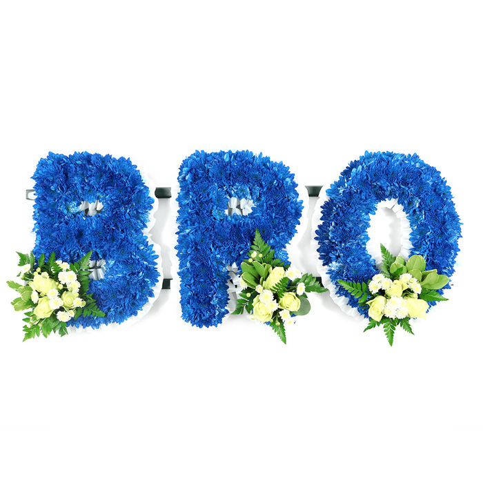 A solemn yet tender three-piece tribute occupies the frame: the word BRO fashioned in large, free-standing capital letters, each letter cloaked in hundreds of royal blue chrysanthemum heads. The blue is rich and saturated, a colour that reads as both calm and steadfast; the tiny florets press together to form an almost velour surface that throws gentle shadows under soft studio light. Near the base of each character sits an elegant spray of pale ivory roses - some tight, like small ovals of cream, others easing open into soft rosettes - accompanied by fine white filler blooms that suggest mini chrysanthemums or baby's breath. These pale accents break the blue with quiet radiance, while mixed foliage - glossy, broader leaves similar to salal or lemon leaf and finer, feathery fern fronds - frames each cluster and gives the design an organic silhouette. A slim white support frame or foam core is visible around the letters' contours and within the loops of the 'B' and 'R' and the hollow of the 'O', indicating the sturdy construction beneath the blooms. The composition is clinical in clarity but warm in intention: a memorial piece meant for wakes, funeral processions, or a chapel setting, evoking peace, sincerity, and familial love. To those in Gerrards Cross and neighbouring communities such as Chalfont St Peter and Denham, this arrangement will read as a respectful, personalised gesture-carefully balanced, quietly dignified, and designed to honour a brother or beloved friend at a service or graveside.