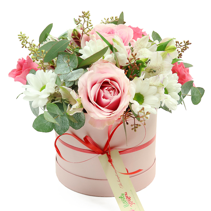This image presents a sophisticated floral hatbox arrangement, styled as though it has just left a boutique florist on Packhorse Road in Gerrards Cross. Centred against a bright white background, the pastel pink, cylindrical hatbox becomes a soft frame for the bouquet spilling from its top. The box has a smooth, matte finish that catches the light gently, giving an impression of quiet luxury. Around its middle sits a narrow satin ribbon in a coral-red shade, tied into a tidy bow at the front. From the knot hangs a slim, rectangular cream tag with elegant green handwriting, implying a personalised dedication ready to accompany the flowers to a home by East Common or a cosy café celebration. Rising from the box, the flowers form a rounded, abundant dome, the blooms carefully layered so no single element feels out of place. At the forefront rests a stunning, fully opened rose in pale pink, its centre tinted with a deeper, more saturated pink that spirals outward through velvety petals. Clustered near it are white chrysanthemum-style daisies, each one boasting a bright yellow-green centre that sparks against the softer tones. Scattered between are white alstroemeria, their elongated petals marked with fine speckles, bringing a graceful, almost featherlike quality. Richer pink and fuchsia blossoms, likely spray roses or lisianthus, introduce depth and a joyful contrast. Around and beneath the blooms, florists' greens weave in and out: rounded silver-green eucalyptus leaves, darker green foliage, and slim stems tipped with fresh buds, adding layers of texture and an airy, natural finish. The overall bouquet appears fresh, balanced, and quietly romantic, ideal for marking milestones, from birthdays to Mother's Day, or as an elegant thank-you delivered across Gerrards Cross.
