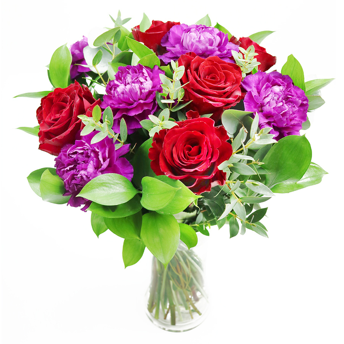This floral arrangement presents a striking yet refined contrast, like an elegant reception setting in Gerrards Cross where tradition and modern style meet with ease. At its heart, velvety red roses open wide, their petals rich and saturated, overlapping in generous layers that evoke timeless romance and deep affection. Nestled among them are lavish purple peonies, each bloom full and rounded, with intricate ruffled petals that give a sense of movement, as if a soft breeze from nearby Austenwood Common had just brushed past. The interplay between the structured roses and the more relaxed peonies creates a visual rhythm that is both classic and fresh. Around these focal flowers, lush greenery extends outward, grounding the arrangement with a sense of natural abundance. The leaves are varied yet harmonious-some broad and glossy, others more delicate-framing the blooms without overwhelming them. Slender branches of eucalyptus weave gently through the greens, their cool, silvery tone adding a note of contemporary elegance and a whisper of invigorating fragrance. The florist has placed the bouquet into a clear glass vase, allowing the stems to be seen in clean, vertical lines, their green and rose-brown tones softened by clear water. The overall effect under soft daylight is almost painterly: reds deepen to wine, purples glow with a jewel-like quality, and the greens provide lively contrast. You can imagine this bouquet on a polished dining table in a Gerrards Cross home, a highlight for an anniversary dinner, a heartfelt birthday surprise, or a special thank-you for someone who loves bold colour with a sophisticated finish. It feels tactile and inviting, as if you could reach out and feel the smooth leaves and plush petals beneath your fingertips.