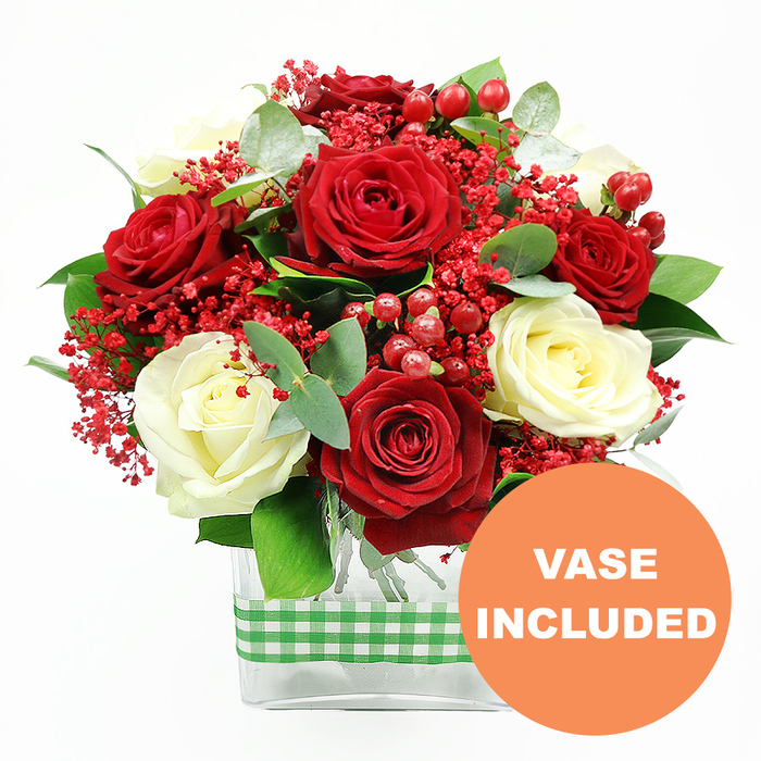 A richly detailed, celebratory bouquet of roses sits centre stage against a bright white background, glowing with the kind of inviting warmth you might find in a Gerrards Cross living room on a winter evening. At the focal point are full-headed red roses, their petals deeply saturated and velvety, curling outward in generous spirals that suggest romance and heartfelt emotion. Interspersed among them, creamy white roses provide a gentle, soothing counterpoint, their ivory tones and slightly tighter petals lending a sense of calm structure to the composition. Weaving delicately between the roses are clouds of tiny, bright red filler blooms, similar to dyed baby's breath, adding a fine, airy texture that softens the transitions between each larger flower. Dotted throughout, glossy red hypericum berries sit like tiny jewels, round and plump, catching light on their smooth skins and reinforcing the festive, almost Christmas-like mood familiar to the streets around Station Road. Surrounding and supporting the flowers, varied green foliage spreads outward: deep green leaves add body and strength, while slender eucalyptus stems introduce a cooler, silvery-green sheen and an imagined fresh, herbal scent. All of this is anchored in a contemporary clear glass rectangular vase, through which the green stems are visible, creating a neat grid of natural lines. Around the waist of the vase, a green and white gingham ribbon is wrapped snugly, its country-style pattern lending a hint of rustic charm that would feel perfectly in place in a home near Chalfont St Peter or by the open spaces of East Common. In the lower right corner of the image, a vivid orange circle displays the words VASE INCLUDED in clean white lettering, highlighting that this arrangement is ready to display on arrival-ideal for anniversaries, Valentine's Day, or simply sending a thoughtful surprise across Gerrards Cross with convenient local flower delivery.