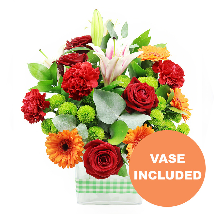 This image shows a lush, densely packed floral arrangement designed in a clean, contemporary style, ideal for brightening a home or office in Gerrards Cross. The bouquet is presented in a clear rectangular glass vase placed against a stark white background, which makes every hue appear more vivid. At the centre and slightly toward the front, several deep crimson red roses bloom with velvety, tightly curled petals, symbolising love and admiration. Intertwined with them are ruffled red-orange carnations, their layered petals creating a soft, cloud-like texture that contrasts beautifully with the roses' more defined form. Scattered throughout the composition, bright orange gerbera daisies stand out with their flat, sunburst petals and dark central disks, evoking warmth and cheer similar to a sunny walk along the tree-lined streets leading to Gerrards Cross station. Between these larger blooms, clusters of small, spherical lime green button chrysanthemums bubble up like beads of colour, adding a playful brightness and helping to draw the eye around the arrangement. Rising gracefully above, white Oriental lilies lend height and elegance; some trumpets are fully open, revealing delicate pink striations and dustings of pollen, while others remain in tight buds that hint at days of continued beauty and subtle fragrance. The flowers are framed by a generous collar of foliage: glossy deep green leaves form a supportive base, while arms of silvery eucalyptus add a soft, cool tone and a slightly wild, natural edge. The clear glass vase exposes the stems inside, giving the design a clean, polished finish. Around the bottom half of the vase, a light green and white gingham band wraps horizontally, suggesting a cosy, country-kitchen charm that would suit many homes near East Common and surrounding areas. In the bottom right corner of the image, a bold circular orange badge partially overlaps the arrangement, carrying the clear white text VASE INCLUDED to show that the glass container is part of the complete floral gift.