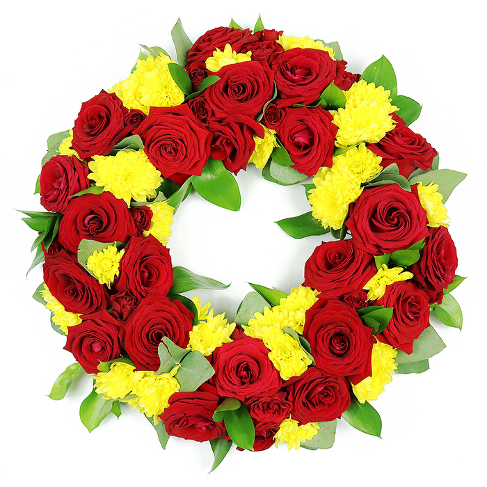 A richly detailed circular wreath designed for funerals or memorials is shown isolated against a stark white background, making the interplay of red, yellow, and green appear almost luminous, as if ready to be placed at a service in Gerrards Cross. The wreath forms a complete ring with a clean, open centre, its outline smooth and consistent, demonstrating the careful craftsmanship of an experienced florist. Dozens of deep red roses are the focal point, their velvety petals layered in tight, classic rose spirals that slowly unfurl towards the outer edges. The shades range from bright, passionate crimson to more shadowed burgundy at the base of some blooms, giving dimension and a sense of quiet depth. Threaded among the roses are plentiful clusters of bright yellow chrysanthemums, likely sprays or small button varieties, their compact, feathery petals creating a soft, cushion-like surface that contrasts with the smoothness of the rose petals. These yellow accents bring a gentle radiance, suggesting comfort and a subtle celebration of life. Supporting the flowers is a thick collar of mixed foliage: broad, slightly waxy green leaves, perhaps salal or ruscus, combined with slimmer, more muted grey-green leaves that echo eucalyptus, some catching the light with a soft sheen, others providing shadowed pockets of calm. The blooms are arranged tightly together so that no gaps show, creating a sense of abundance and completeness often requested for services in and around Gerrards Cross, from quiet family gatherings to more formal commemorations. The white background and even lighting emphasise every texture and nuance of colour, turning the wreath into a powerful visual tribute that speaks of love, respect, remembrance, and enduring connection.