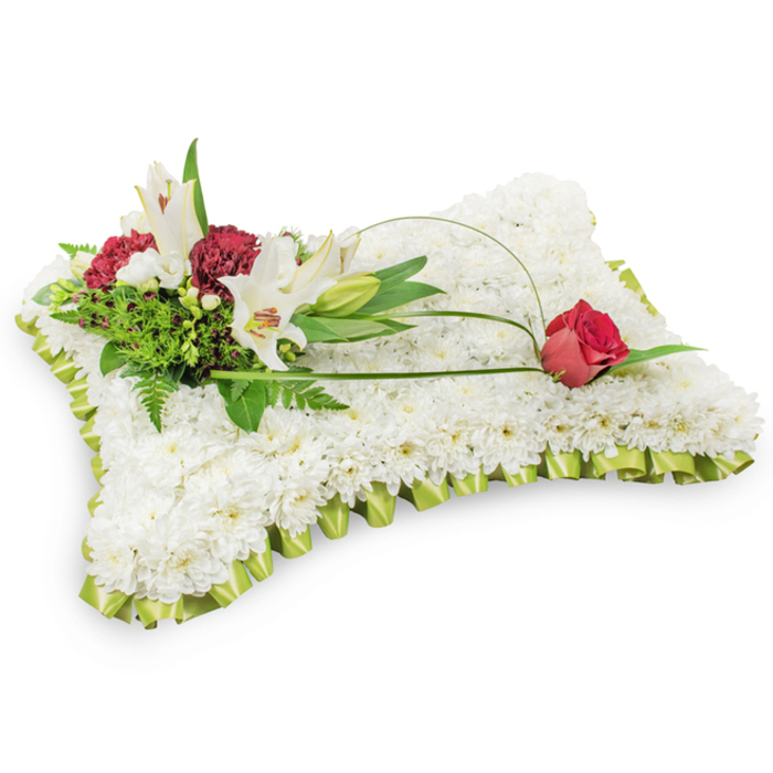 Photographed on a simple white field, this classic funeral pillow is a carefully composed expression of solace. The main rectangular form is entirely cloaked in hundreds of tiny white chrysanthemums, each bloom placed to create a uniform, cushion-like surface that reads as both refined and tender. A neat, pleated border of pale lime-green satin surrounds the base, its soft folds catching gentle highlights and giving the cushion a refined, almost textile-like finish. On the upper left a naturalistic cluster rises: two elegant white Oriental lilies open to reveal their soft yellow anthers while a nearby lily bud curves upward with quiet promise. Interspersed among the lilies are rich burgundy spray carnations, whose textured petals bring warmth and a dignified counterpoint to the white. Layered behind these blooms, varied green foliage - including dainty fern fronds and broader leaves - adds shadowed depth and a living quality to the group. From that cluster, a slim green tendril of bear grass or salal arcs across the chrysanthemum field, drawing the eye to a single deep red rosebud resting on the upper right; the rose's velvety surface and closely held petals embody respect and remembrance. The arrangement evokes a sense of calm and purity, the scent understated, the composition balanced and intentional. It is a soothing tribute suited to memorial services and wakes in Gerrards Cross and nearby Beaconsfield or Denham, designed to bring comfort to those grieving while honouring the departed with classic floral symbolism.