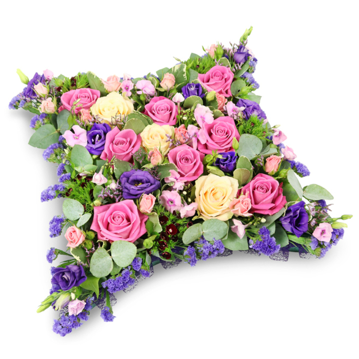 This image shows an elegant floral cushion tribute, shaped like a soft, rectangular pillow with rounded corners, carefully composed to convey comfort and loving remembrance for a farewell service in Gerrards Cross. The entire surface is densely packed with fresh-cut flowers, giving it a plush, padded look that feels gentle and inviting, as if it could cradle cherished memories. Large roses in shades of vibrant fuchsia, warm mid-pink, and creamy butter-yellow form the main body of the design, their petals tightly layered in some blooms and more open and full in others, revealing soft, swirling hearts. Intertwined among them, luxurious lisianthus flowers in a rich royal purple and slightly lighter violet tones create a striking contrast, their velvety petals and softly ruffled edges giving the arrangement a refined, almost regal air. Tiny lavender-hued filler blossoms, resembling statice or limonium, are scattered throughout, adding an airy, lace-like texture that softens the transitions between the bigger flowers. Lush green foliage provides a cool, tranquil foundation: rounded, silvery eucalyptus leaves bring a gentle, powdery green, while deeper green, fern-like strands stretch delicately between blooms, adding shadows and depth. The clean white background and subtle shadow beneath the arrangement make the colours appear especially vivid, highlighting the craftsmanship and balance of the tribute. The overall impression is one of serenity, compassion, and respect, perfectly suited to a funeral, cremation, or graveside service. Crafted with the care you would expect from a trusted Gerrards Cross florist, this floral pillow feels like a heartfelt, tangible expression of sympathy, the kind of tribute that might rest beside a photo frame or candle at a memorial gathering near West Common or the local parish church.