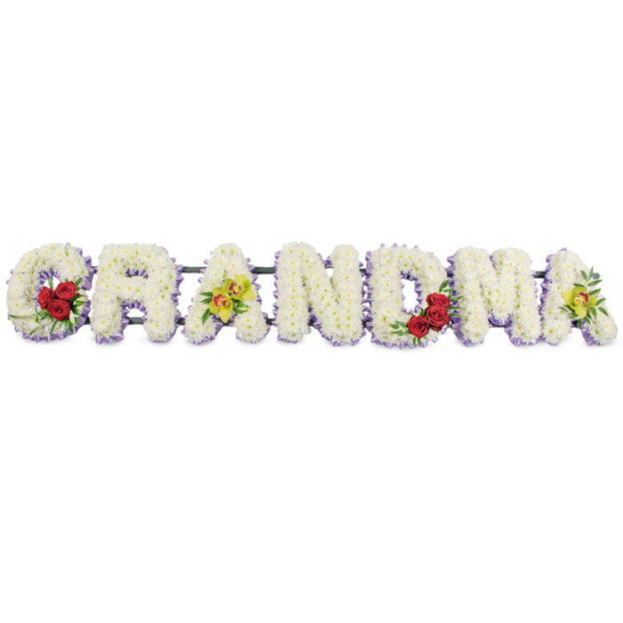 A dignified floral arrangement spells out the word GRANDMA in large, sculpted capital letters, stretching horizontally across the frame against a pure white background that makes every shade of petal and foliage stand out clearly. Each letter is meticulously covered in tiny white chrysanthemums or similar button blooms, placed so closely together that they form a smooth, padded surface, reminiscent of soft snow yet full of life and texture. At the base of each letter, a fine trim of pale lavender flowers peeks through, running like a soft lilac shadow along the bottom edge and giving the tribute subtle definition and depth. The florist has introduced small, meaningful bursts of colour to personalise the design. On the lower curve of the 'G', three deep red roses, surrounded by polished dark green leaves, rest together, their rich tone suggesting devotion and heartfelt thanks. The first 'A' is adorned with two chartreuse cymbidium orchids lying gracefully across its centre, their yellow-green petals and speckled reddish throats providing a contemporary, elegant contrast to the white base. The 'R', 'N', and 'D' remain unembellished, a calm run of white and lavender that feels serene and contemplative. The 'M' is accented on its lower right side with another trio of red roses, echoing the love expressed in the opening letter, while the final 'A' mirrors the first with two more orchids, creating a balanced, harmonious finish. All the letters are supported by a slim dark green metal framework that connects them subtly along the top, designed to sit securely on stands or an easel during a service in Gerrards Cross, perhaps in a local church or at the crematorium. The arrangement communicates quiet respect and affection, a gentle way for a family to say farewell to a much-loved grandmother while surrounding her memory with beauty.