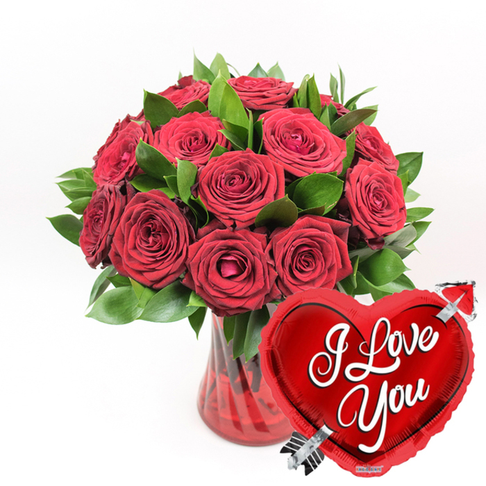 A richly romantic arrangement of deep red roses appears as if it has just been placed on a pristine white tabletop in a Gerrards Cross home, perhaps overlooking the leafy calm near West Common. The bouquet features approximately a dozen or more roses, each bloom showcasing layers of velvety petals in a luxurious crimson hue. Some flowers are fully open, revealing their intricate centres, while others are still unfurling, their petals forming gentle spirals that suggest tenderness and anticipation. Interwoven among the roses is a generous amount of vivid green foliage, the leaves broad, glossy, and full of life, wrapping the bouquet in a natural, textural frame that heightens the drama of the red. These stems are gathered into a stylish ruby-red glass vase with subtle fluted lines that run vertically along its surface. The vase is translucent, allowing glimpses of the stems inside, and the glass catches the light in delicate highlights, much like the reflections in a shopfront window along Station Road. In the lower right foreground, slightly overlapping the vase, a shiny, metallic red heart-shaped balloon tilts toward the viewer. Its surface gleams with reflections, giving the balloon a three-dimensional, almost buoyant presence. Across its centre, the message I Love You is written in bold, elegant white script, clearly legible and emotionally direct. A silver Cupid-style arrow seems to pierce the heart from the lower left to the upper right, adding a playful, storybook touch. The entire scene is set against a crisp white background, intensifying the contrast between the lush greens, rich reds, and gleaming silver. This image captures the essence of a grand romantic gesture, perfect for same-day delivery from Flower Delivery Gerrards Cross to mark Valentine's Day, an engagement celebration, or a meaningful anniversary shared quietly at home.