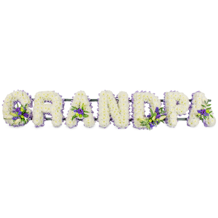 The image shows a carefully composed funeral flower tribute spelling GRANDPA in strong, capital letters, laid out in a steady horizontal row as though ready to be placed beside a coffin or at a memorial near Gerrards Cross Common. Each letter is built on a sturdy base, then entirely cloaked in a rich covering of small white flowers-mainly chrysanthemums or carnations-set so tightly that the surface appears almost like a soft, snowy cushion. The repetitive shapes of the petals create a calming rhythm, while the creamy white colour suggests innocence, reverence, and a peaceful passing. Here and there, the serenity of the white is gently interrupted by pockets of deeper colour: clusters of purple and lavender blooms, possibly lisianthus or similar seasonal flowers, are nestled within the curve of the G, centred in the first A, scattered thoughtfully through the D and the P, and lending a final grace note to the last A. These accents, supported by sprigs of fresh green foliage, add movement and depth, like small bouquets tucked into each letter. Beneath the flowers, a narrow dark green frame or rail runs almost invisibly along the base, linking the individual letters so they stand together as one message of love. The arrangement feels personal and local, the kind of bespoke piece a Gerrards Cross family might order when gathering after a service to share memories of their grandfather. Its blend of pure white tones and serene purple highlights evokes a quiet sense of comfort, making this tribute not just a name in flowers, but a soft, visual echo of a life cherished and remembered.