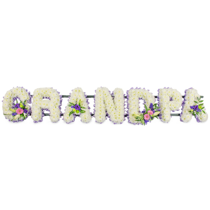 In this image, the word GRANDPA appears as a full floral display, each letter proudly shaped from fresh blooms and laid out horizontally across a stark white background that makes the design seem to float. The letters are robust yet soft in appearance, completely covered in a dense layer of white chrysanthemums or similar small, multi-flowered heads, giving a plush, almost quilted surface that suggests comfort and tenderness. Along the outer edge of every letter runs a narrow band of tiny lilac and light purple flowers, possibly statice or asters, tracing each curve and corner with a subtle line of colour, like a pastel frame that adds clarity without overwhelming the overall whiteness. Several letters hold deeper accents that draw the eye gently along the tribute: the 'G' carries a small spray of pink and fuchsia flowers mixed with a slender white lily and rich green foliage; the first 'A' and final 'A' are lightly adorned with compact purple florets and dark leaves; the 'D' features a more prominent grouping that echoes the 'G', again combining graceful white lily petals with cheerful pinks and fresh greenery, sometimes joined by bright green button-style blooms for extra texture. All the letters are supported by a discreet dark green bar or frame that runs behind them, suggesting this name tribute is intended to stand upright, perhaps at the front of a funeral service in Gerrards Cross or beside the resting place in a local cemetery. The overall effect is one of serene remembrance and personal affection, a customised floral farewell for a cherished grandfather whose presence is lovingly spelled out in flowers.