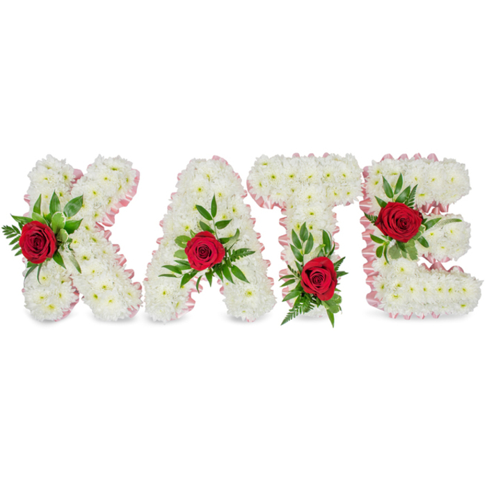 A large, bespoke floral name tribute presents the letters K A T E in bold three-dimensional block form, each letter built from a dense, uniform covering of small white chrysanthemums that give a plush, almost cloud-like texture set against a stark white background. A light pink satin ribbon outlines every contour and corner, neat and pleated, softening the geometry and lending a gentle blush along the edges. Each letter carries a single, striking deep crimson rose as its focal point, accompanied by a generous spray of green foliage - broad, glossy leaves and slender ferny fronds that frame the roses and add layered depth. The floral accents are positioned with precision: the rose on the K resides on the lower portion of its left vertical stroke; on the A the rose is placed centrally at the triangular peak; the T's rose is tucked at the base of the left side of its horizontal crossbar; while the E's rose sits on its middle horizontal bar. The arrangement is photographed in soft, diffuse light that highlights petal texture, the satin sheen of the ribbon and the contrast between the white chrysanthemum mass and the deep red roses. This composition is typical of dignified funeral and memorial lettering - a design I regularly prepare for services in and around Gerrards Cross, including families from Packhorse Road, Beaconsfield and neighbouring Denham - offering a quiet, classical expression of purity, love and remembrance executed with calm precision.