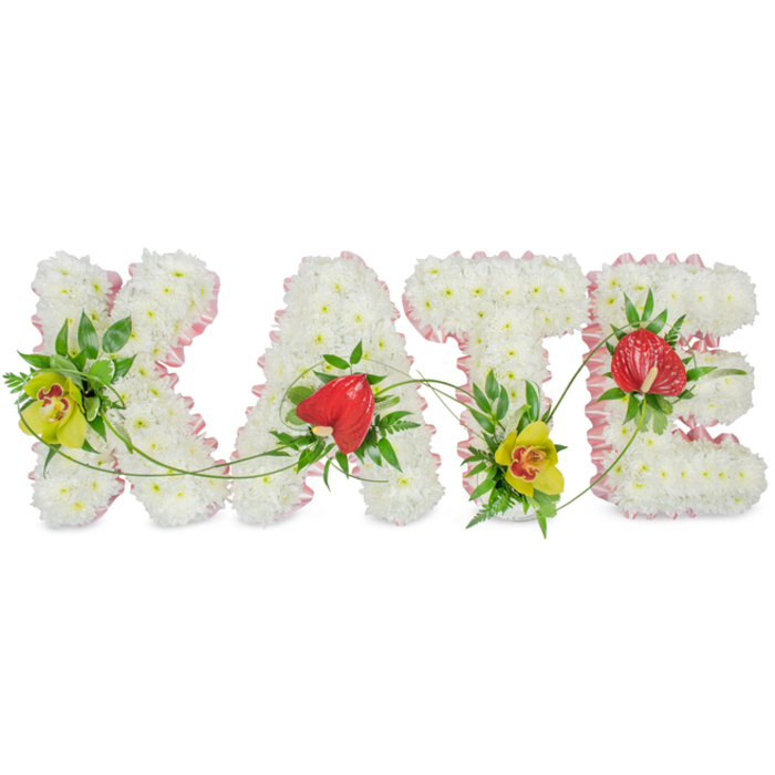 The image shows a carefully handcrafted funeral flower tribute spelling out the name KATE in four bold floral letters, placed against a clean white background so the colours and shapes are perfectly clear. Every letter is built entirely from clusters of small white chrysanthemums, packed so closely that they form a plush, almost cloud-like texture, giving the piece a sense of purity and calm remembrance. Running all around the outside edge of each capital letter is a narrow, light pink ribbon, folded and softly ruffled, which frames the white blooms with a gentle blush of colour. This subtle trim adds a feeling of tenderness and refinement, ideal for a memorial or church service in Gerrards Cross. Carefully positioned accents bring the name to life: on the K and T, lime-yellow cymbidium orchids with speckled reddish-brown centres provide striking focal points, their glossy petals and intricate markings adding an elegant, exotic note. The A and E are adorned with deep red, heart-shaped anthuriums, their smooth, waxy surfaces contrasting beautifully with the fluffy chrysanthemums and suggesting deep affection and lasting love. Around these statement flowers, rich green foliage is tucked in - broad leaves, ferns, and small sprigs - adding depth, structure, and a natural garden feel. Slender, looping green vines trail gracefully across and between the letters, visually tying the whole name together as one flowing tribute. This bespoke arrangement, ideal for same-day funeral flower delivery to crematoriums and churches around Gerrards Cross and nearby villages, expresses a personal farewell to Kate with dignity, warmth, and local florist expertise.