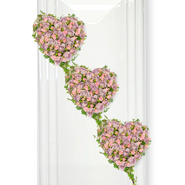 A sophisticated three-heart funeral arrangement fills the frame, each heart shaped entirely from a tapestry of delicate miniature roses. Positioned in a subtle diagonal cascade from top left to bottom right, the hearts create a feeling of continuity, as though love is flowing gently from one to the next. The roses themselves are predominantly a soft, dusty lavender, interspersed with lighter pastel pink blooms that brighten the design and add gentle contrast. Each bloom is placed closely beside the next, so that no space shows between petals, resulting in a rich, almost quilted effect that feels both luxurious and comforting. The petals are slightly ruffled at the edges, suggesting freshness and softness, and the mix of buds and open roses lends a natural sense of life and movement. Framing and linking the hearts, bright green ivy vines weave around the contours, their small, glossy leaves outlining each heart shape with precision. These trailing strands connect the hearts vertically, like a living garland, and bring a relaxed, garden-like charm reminiscent of the leafy paths and quiet corners around West Common and the surrounding lanes of Gerrards Cross. The arrangement is photographed against a brilliant white background, with subtle translucent vertical elements on the left that hint at glass or polished paneling, adding a refined, almost gallery-like atmosphere. The overall effect is serene and deeply tender, making this triple-heart design a fitting tribute for a funeral, memorial, or celebration of life, especially when you wish to convey enduring love between partners, siblings, or generations with flowers arranged by an experienced local Gerrards Cross florist.