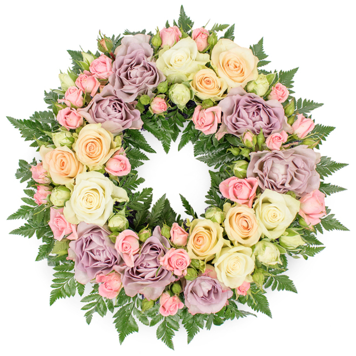 Set against a plain white backdrop, a sumptuous memorial wreath of roses and fern foliage forms a complete, unbroken circle that feels both serene and deeply personal. The design is tightly built, with each bloom nestled closely to its neighbour so the eye moves in a soft, continuous sweep around the ring. Cream and ivory roses dominate, many at full, luxurious openness with layers of petals unfurling from a tight heart, giving a sense of fullness and comfort. Interspersed among these are gentle pastel shades: pale peach roses with a warm glow, buttery yellow blooms that brighten the palette without overpowering it, and distinctive dusty lavender or mauve roses that lend a refined, almost heirloom character. Clusters of delicate pink spray roses are tucked into gaps, some still in bud, others just beginning to open, adding smaller points of interest and suggesting the progression of life from youth to maturity. Between and beneath all these blossoms, finely cut dark green fern fronds weave through the structure, their feathery tips just visible, outlining the wreath and providing a deep, verdant counterpoint that makes the lighter petals stand out. The hollow centre of the wreath remains clear, emphasising the circular symbolism of eternity and the ongoing nature of love, even after loss. This is the kind of carefully composed tribute one might see at a funeral service or graveside in Gerrards Cross, thoughtfully prepared by a local florist who understands how soft colour, lush texture, and meticulous balance can bring a quiet sense of solace to families gathered to say goodbye.