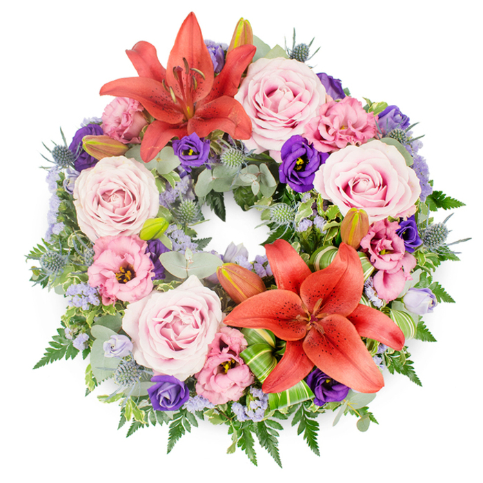 Set against a bright white background, this round funeral wreath appears almost like a painted garland brought to life, every petal placed with care. The overall shape is a perfect circle, full and lush, with no gaps, giving a reassuring sense of completeness and continuity. Two large Asiatic lilies in deep coral-orange dominate the upper and lower sections; their starry, flared petals and clearly visible stamens provide bold accents that stand out against the softer surrounding pastels. Nestled around them are numerous roses in shades of soft blush to lavender-pink, their layered, spiralling centres fully open in some blooms and gently cupped in others, suggesting a tender progression of time and memory. Interlaced throughout the wreath are lisianthus (eustoma) in two main tones: delicate pale pink-lilac and rich, velvety purple. Their ruffled edges and cup-shaped flowers introduce a gently romantic, almost textile-like texture, as if the flowers were sewn together in a tapestry of remembrance. Fine clusters of lavender-blue filler flowers, likely statice or limonium, peek out in small clouds, creating a misty veil that softens transitions between the more prominent blooms. The greenery is abundant and varied: rounded, silvery eucalyptus leaves catch the light; fine fern fronds extend in feathery arcs; and striking blue-grey eryngium thistles add spiky, architectural accents that echo the wild beauty of the Buckinghamshire countryside near Gerrards Cross. Several firm, green lily buds are tucked discreetly among the foliage, suggesting life and hope continuing beyond loss. Artfully folded bright green leaves near the lower lily introduce a modern note that speaks to skilled local floristry. This wreath feels perfectly suited to a thoughtful farewell, whether delivered to a quiet family service or a larger gathering within the Gerrards Cross community.