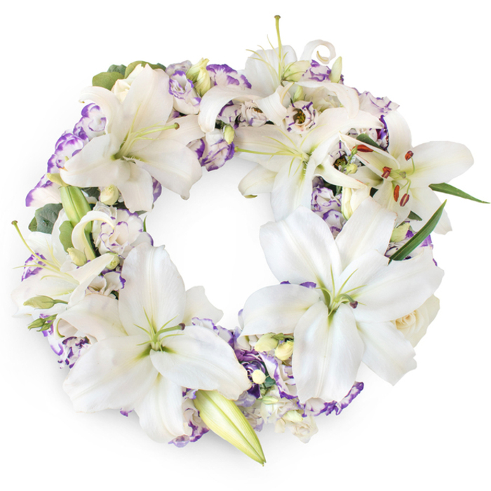 An impeccably arranged circular floral wreath is displayed on a crisp white background, its complete ring of flowers forming a soft halo-like shape. The wreath is generously adorned with large white Oriental lilies, which serve as the central focus of the design. Their wide, satiny petals open outwards in graceful arcs, revealing pale green centres and elongated stamens dusted with fine, warm-toned pollen. Some lilies are in full, majestic bloom, while others remain partially closed or held in elegant, tapered buds, creating visual layers and a natural, organic rhythm. Interwoven among these signature blooms are clusters of Lisianthus, their petals creamy white with gently frilled edges, each petal traced with delicate lilac and muted purple tones. This subtle colouring introduces a tender contrast, never overwhelming but instead echoing the quiet emotions of remembrance. Occasional small buds hint at new life, adding a touch of hope to the composition. Beneath and between the flowers, sprigs of rich green foliage and cool-toned, eucalyptus-like leaves emerge, providing structure, shadow, and a sense of depth without distracting from the purity of the white and purple palette. The overall mood is one of calm, reverent beauty, perfectly suited to a farewell in Gerrards Cross, whether at a church on Packhorse Road or a private gathering at a local chapel. This sympathy wreath feels both traditional and heartfelt, the continuous circle symbolising everlasting love, the lilies evoking peace and the soul's journey, and the soft purple Lisianthus whispering of dignity and gentle sorrow. It is an arrangement that quietly supports mourners, honouring a cherished life with understated grace.