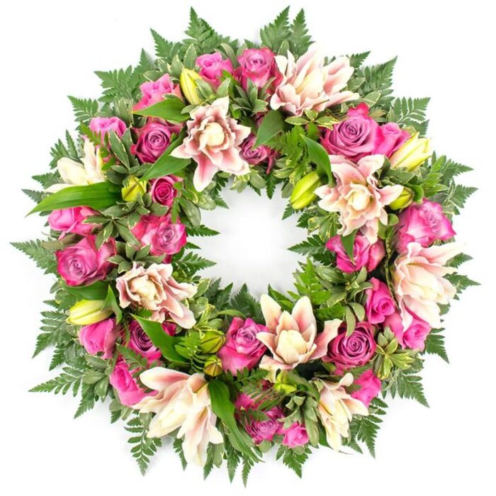 This image shows an opulent sympathy wreath captured from above, perfectly round and densely filled so that the entire circle appears rich and complete. The florist has worked with a harmonious blend of pink and cream flowers, allowing the colours to flow gently around the ring rather than in strict bands. Rose heads dominate the design, their tones moving from strong magenta to lively fuchsia and into delicate blush pink. Some roses sit high and proud, petals cupped closely as if they have just begun to open, while others are fully expanded, their layered centres revealing a soft spiral that draws the eye inward. Nestled among these roses are double lilies, their multiple petals lending a luxurious, ruffled texture. The lilies show soft gradients of colour: creamy white at the base, then warming into pale peach and blush towards the tips, with occasional warm pink highlights that echo the surrounding roses. A scattering of plump, lime-green lily buds rises from the greenery, adding freshness and a sense of life continuing. The floral ring is supported and framed by generous foliage. Fern fronds in deep forest green radiate along the edges, their finely divided leaflets creating a feathery, almost mist-like border. Behind and between them, broader leaves in various greens, some lightly variegated, reinforce the circular shape and hide the underlying frame. The lighting is bright and neutral, so each nuance of colour and texture is visible against the stark white background, much as the wreath might stand out when delivered to a peaceful chapel of rest or a family home off Packhorse Road or around Bulstrode Way in Gerrards Cross. The composition feels carefully balanced and deeply respectful, the unbroken circle symbolising eternity and ongoing love in times of farewell.