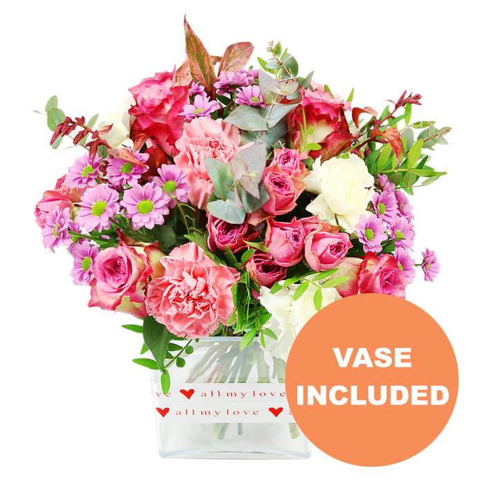 This image showcases a richly detailed mixed bouquet that practically spills out of its clear glass vase, set against a spotless white background that makes every colour stand out. At first glance, your eye is drawn to the large, luxurious roses in varying shades of pink: some are intense fuchsia and magenta, others are more muted blush tones, and a few appear to have slightly deeper edges that give a softly shaded effect. Entwined among them are full, ruffled carnations in coral and peach-pink, their densely layered petals adding volume and a tactile, feathery look. Punctuating the arrangement are daisy-like chrysanthemums in lavender and soft purple, each bloom centred by a bright green disc that creates a lively contrast to the surrounding warm hues. Creamy white flowers, delicately placed between the bolder blooms, lend an airy, elegant touch and stop the palette from feeling too heavy. The bouquet feels lush and generous thanks to the foliage: rounded eucalyptus leaves in a silvery-green tone mingle with deeper green leaves, and a few slender stems of reddish-brown foliage introduce just a hint of rustic, romantic depth. The flowers are arranged in a minimalist, square glass vase whose clear sides show the neatly trimmed stems within, suggesting the kind of professional finish you'd expect from a local Gerrards Cross florist preparing an order for a home off Bulstrode Way or near the Memorial Centre. Wrapped around the base of the vase is a white decorative band featuring repeating red heart motifs and the loving phrase all my love in flowing script. At the lower right corner of the photograph, an orange circular badge with bold white letters states VASE INCLUDED, highlighting that this arrangement arrives as a complete, ready-to-enjoy gift for occasions like Mother's Day, anniversaries, birthdays, or simply to brighten a quiet afternoon in Gerrards Cross.