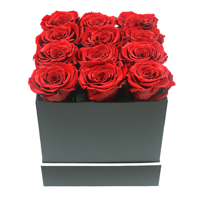 This image captures a sophisticated rose arrangement at eye level, as if the box has just been set down on a table in a calm Gerrards Cross living room. A perfectly square, matte black gift box holds twelve red roses in a disciplined 3x4 grid, each bloom identical in size and stage of opening. The roses are fully unfurled yet still tight at their centres, creating a pattern of spiralling petals that appear richly textured and velvety to the touch. Their hue is a deep, romantic crimson, with shadowed undertones that drift toward burgundy in the folds, contrasting powerfully with the pale, seamless white background. The box itself is sleek, its edges sharp and geometry uncompromising, with no shine to distract from the flowers. Near its base runs a single narrow white band, almost like a minimalist frame, giving the design a polished, boutique feel reminiscent of high-end gifts exchanged around Gerrards Cross, from quiet roads near the Memorial Centre to elegant homes off Oxford Road. The lighting is soft and diffuse, without any harsh glare, so the petals seem to soak up the light, and faint, gentle shadows beneath the box hint at depth and dimension. There are no other props or decorative elements - no greenery, ribbons, or text - just the pure statement of ruby red against black and white. The composition suggests the kind of preserved or 'eternal' roses that remain beautiful for months, ideal for long-lasting romantic gestures, celebratory keepsakes, or sophisticated décor that can sit gracefully on a bedside table or coffee table in Gerrards Cross, quietly radiating love and intention.
