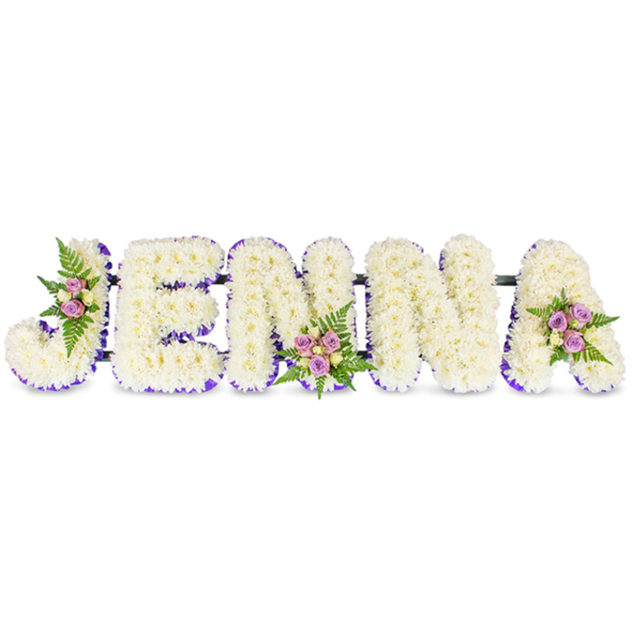 A lovingly crafted memorial spells JENNA in five substantial, three-dimensional letters set cleanly against a white backdrop, each letter densely composed of creamy white chrysanthemums or similar petite florets that produce a soft, cloud-like texture. The arrangement's quiet palette is punctuated by a vivid, deep purple trim or ribbon that traces the inner edges and bases of the letters, lending structure and a dignified contrast to the pale blooms. Three distinct accent posies are arranged by hand: one nestles into the upper left curve of the J, another is placed at the lower centre of the second N, and a final cluster rests on the upper right of the A. Each cluster contains delicate blush-pink and lavender spray roses, complemented by a few lighter cream or pale yellow roses and framed by fresh, dark-green fern fronds that add graceful movement and a whisper of woodland scent. Slim, dark support bars run subtly behind the letters, joining them into a single, stable composition intended for display at wakes, funerals, or a celebration of life. The soft studio lighting emphasizes the tactile quality of the petals and the gentle contrast between white and purple, evoking calm and respectful remembrance. This bespoke name tribute would be at home in the chapels and churches serving Gerrards Cross and the surrounding villages of Beaconsfield and Chalfont St Peter, offering families a personal and heartfelt floral statement to honour a loved one with quiet beauty and local craftsmanship.