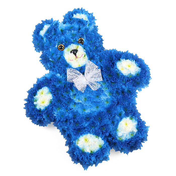 This image shows a beautifully sculpted floral teddy bear laying slightly on the diagonal against a pure white background, as though cradled in light. Its entire form is built from fresh blossoms, with royal blue flowers packed so closely they create a soft, almost cloud-like texture over the rounded head, torso, and limbs. The intense blue is steady and saturated, yet softened by the natural variation of petals, with some catching highlights and others falling into subtle shade, which gives the bear depth and gentle curves. Cream to pale yellow blooms form the inner parts of the bear's ears, a rounded snout at the centre of its face, and four neat oval pads on its paws, like the stitched fabric details on a favourite childhood toy. These lighter accents bring warmth and definition, guiding the eye from ear to face to feet. The expression is completed with two shiny, dark brown eyes, each with a black pupil that reflects a small glint, a simple black triangle nose, and a slight curved line beneath, together suggesting a quiet, reassuring smile. Around the neck, a fine white mesh bow is tied with care, its translucent loops and tails softening the blue further and adding an air of innocence and ceremony. The overall impression is of tenderness and remembrance, perfectly suited as a funeral tribute or memorial design for a child or baby boy. Created in the style often requested by families around Gerrards Cross and nearby Chalfont St Peter, this floral teddy bear offers a gentle, tactile way to express love, sorrow, and an enduring wish to hold someone close in memory.
