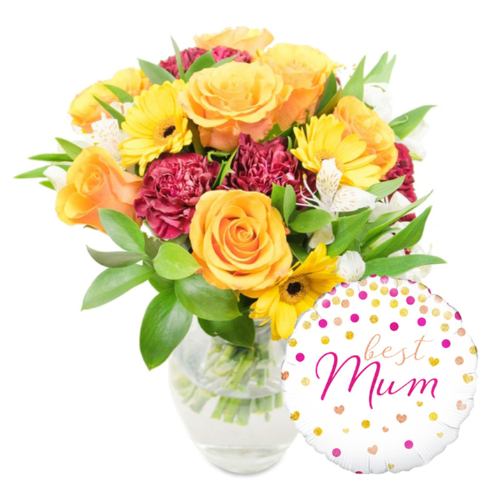 A vivid, heart-warming floral gift set sits against a pristine white background: a generous bouquet in a clear, gently fluted glass vase occupies the centre-left, while a glossy round balloon rests at the lower right, slightly overlapping the vase. The bouquet is built on warm, uplifting tones-ample orange roses with tight, spiralled petals create a soft, luminous centre, complemented by bright yellow gerbera daisies that bring a sunny energy and clear, dark centres for contrast. Deep red-burgundy carnations, their densely ruffled petals like tiny velvet cushions, add weight and romance to the grouping. Accents of white alstroemeria, with narrow, freckled petals, introduce delicate, refined points that catch the light and give the eye places to rest. A full complement of fresh greenery-smooth broad leaves and airy, linear foliage-wraps the blooms and is visible through the transparent vase, where cut stems stand in clear water, conveying recent preparation and freshness. The round foil balloon is white with playful gold and fuchsia confetti-like dots and little hearts; across its face best Mum is written in an elegant fuchsia script, the words curving with affectionate emphasis. The overall mood is celebratory yet intimate, evoking soft morning sunshine on Gerrards Cross High Street and the quiet pleasure of giving. This arrangement is a popular thoughtful choice for Mother's Day or birthday deliveries around Chalfont and the surrounding local area.