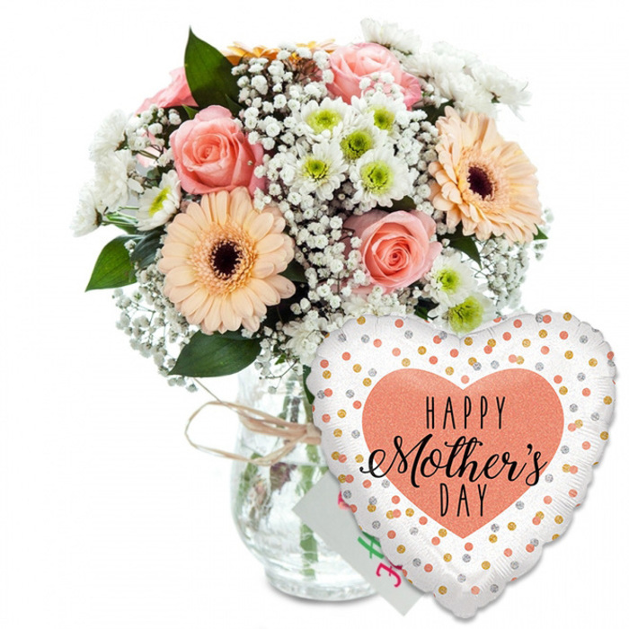 This image shows a thoughtfully arranged Mother's Day gift set, photographed against a bright, uncluttered white background so every detail of the flowers and balloon is easy to imagine. On the left, a clear glass vase holds a rounded bouquet of mixed blooms, the water line visible through the glass and the neck gently wrapped with a casual bow of natural twine, giving the piece a touch of rustic charm you might associate with a small independent florist just off the centre of Gerrards Cross. The bouquet is dominated by several peach and blush-pink roses, their petals open and layered, creating soft, romantic swirls of colour. Beside them, two or three striking gerbera daisies in warm peach tones stand out, each flower face perfectly round with a dark brown, almost black centre, like a polished button anchoring the bloom. These bold gerberas are softened by the addition of airy white baby's breath, whose tiny blossoms appear like a delicate mist drifting between the larger flowers. Smaller white chrysanthemums with pale green centres nestle throughout the bouquet, building fullness and giving a daisy-like freshness that keeps the composition light rather than heavy. A scatter of rich green foliage supports and surrounds the blooms, adding depth, shadows, and the sense of something freshly gathered. To the right of the vase, partly hiding its base, a heart-shaped foil balloon leans cheerfully towards the flowers. The balloon is crisp white, sprinkled across with metallic gold and silver polka dots that suggest confetti caught mid-air. In the centre, a large peach-coloured heart frames the words HAPPY Mother's DAY in contrasting black fonts, with Mother's rendered in a graceful script that feels handwritten. Just behind the balloon, the corner of a small white greeting card peeks out, reinforcing the idea of a complete, ready-to-give Mother's Day surprise, ideal for doorstep delivery to homes around Gerrards Cross and the surrounding villages.