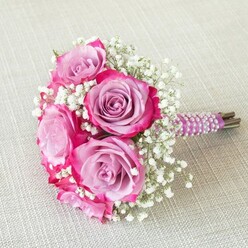 Against a backdrop of softly textured, pale linen-evocative of a refined table at a Gerrards Cross celebration-a vibrant wedding bouquet radiates romance. The heart of the design lies in five glorious roses: their petals unveil a gentle gradient, transitioning from serene lavender at the centre to bright, lively fuchsia brushing each outer edge, creating a visual depth that feels almost watercolor-like. Wisps of snowy white gypsophila are interspersed between the roses, lending a cloud-like softness and a subtle, airy volume throughout. The stems are clasped tightly within a band of opulent, deep purple satin ribbon, whose surface glows as luminous ivory pearls spiral with precision and care, mirroring the refined detailing cherished by many local brides. Hints of the bouquet's natural stems can be seen beneath the ribbon, providing a fresh, just-picked touch. Soft, natural daylight caresses the flowers, drawing out their hues and enhancing the bouquet's layered textures. Whether destined for a wedding at a Gerrards Cross country venue or as a gift to commemorate a milestone anniversary, this arrangement reflects the dedication, artistry, and local pride of skilled florists crafting memories in their community.