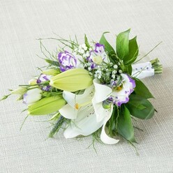 An elegant, elongated bouquet lies diagonally across a pale, textured beige cloth, the kind of simple, natural backdrop you might find in a calm Gerrards Cross home preparing for a wedding morning. Dominating the composition are several large white lilies, fully open and serene, their silky petals gently arching outward around prominent yellow stamens dusted with warm pollen. Faint speckles of reddish-brown decorate the inner petals, adding a touch of quiet complexity to each blossom. Nestled alongside these open flowers are sleek, chartreuse-green lily buds clustered along the stems, their tapered shapes suggesting blooms still to come, giving the bouquet a sense of unfolding time. Interwoven throughout are Lisianthus blooms in white and rich purple, with ruffled, almost frilly petals; some flowers are edged in bold violet, others soften into lilac tones, creating a painterly blend of purple hues. Delicate sprays of white baby's breath float between the larger blooms, their tiny blossoms forming a soft, cloud-like veil that lightens the overall look. A foundation of lush greenery anchors the piece, with broad, glossy leaves offering structure and depth, while slender, wispy foliage and a few spikier green accents add movement and a natural, freshly gathered feeling, as though picked from a garden near West Common. The stems are carefully bound in a smooth white satin ribbon, accented with subtle pearl-style beads that catch the light in a gentle shimmer, hinting at bridal elegance and special occasions. The lighting is soft and even, emphasising the crisp whites, vibrant purples and various greens, and giving the bouquet an aura of calm celebration, perfect for a ceremony, anniversary, or refined gift delivered locally around Gerrards Cross.