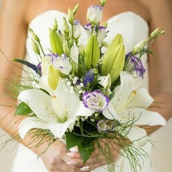 With a touch of romance and a flourish of expert skill, this handcrafted bridal bouquet echoes the quintessential charm of weddings in Gerrards Cross-imagine the gentle hum of St. James's Church or celebrations along Packhorse Road. The bouquet's design is anchored by opulent white Oriental lilies, their ample petals gleaming against the bride's classic white dress, each petal catching natural light in creamy, soft waves reminiscent of fresh snowfall. Scattered artfully among them are lisianthus blooms-pristine white blossoms with distinctly ruffled lavender-violet fringes, their presence adding both texture and whimsy. Wispy fern fronds and sprays of baby's breath grace the bouquet with movement and a cloud-like softness, a nod to the dewy gardens found in Gerrards Cross at dawn. Deep purple accents, subtle yet striking, punctuate the arrangement, inviting the eye to linger on each exquisite detail. Every stem feels thoughtfully positioned, as if gathered from the fields that ring Gerrards Cross, conveying not just freshness but a sense of place and belonging. The scent of lilies mingles with verdant foliage, conjuring memories of spring and new beginnings-making this bouquet a cherished accessory on a day filled with hope, tenderness, and enduring love.