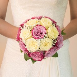 Nestled within the hands of a bride from Gerrards Cross, this resplendent bridal bouquet radiates both expert craftsmanship and local character. Creamy, pale yellow roses are clustered tightly at the heart, their velvety petals catching a soft indoor glow, while striking fuchsia pink roses ring them with notes of lavender and mauve-each shade echoing the subtle romance of a springtime stroll through West Common or along Packhorse Road. Wisps of airy white baby's breath thread between blooms, imparting an ethereal, cloud-like lightness, offset by the occasional dark green foliage that peeks from beneath. The bride's lightly tanned arms frame the bouquet, her fingers gently supporting its round, balanced shape at waist level against the backdrop of her classic ivory wedding gown. The upper dress is adorned with swirling lace, evoking timeless elegance and hinting at cherished family traditions, while the skirt flows in soft, unbroken lines of smooth fabric. Gentle, even light plays across the scene, emphasizing the freshness of petals and the interplay of textures. The softly blurred, neutral background keeps the vision pure and uncluttered, drawing all focus to the bouquet and the emotions it encircles: anticipation, joy, and the promise of new beginnings. Crafted for a bride whose story unfolds in Gerrards Cross, this bouquet lingers in the heart, destined to become a cherished memory.