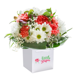 The bouquet captured here exudes the fresh vibrance of Gerrards Cross in full bloom, as if gathered after a morning stroll along West Common's dew-tipped grass. A brilliant combination of coral, peach, and ruby-red carnations forms the heart of the arrangement, their intricately ruffled petals resembling spun silk and imbuing the bouquet with warmth and texture. Gleaming nearby, clusters of white daisies with sun-bright chartreuse centers bring a gentle cheer reminiscent of wildflower meadows nearby. Wisps of delicate white baby's breath float like soft mist, weaving between the larger blooms and softening the bouquet with a cloud-like touch. A few closed buds with hints of purple deepen the visual intrigue, suggesting new beginnings or the anticipation of a special day. The flowers rest within a lush embrace of deep green, glossy leaves, carefully fanned out by hand to provide a full and natural frame. The square white gift bag, adjusted for easy carrying and thoughtfully presented on a pure white background, underscores the arrangement's premium freshness-perfect for birthdays, anniversaries, or surprise deliveries across Gerrards Cross. The elegant composition, expertly arranged, evokes joy, gratitude, and the comforting presence of florals crafted by a florist rooted in local traditions. This bouquet feels like the promise of celebration, ready for same-day delivery whether it's a milestone in Fulmer or a cherished gathering on Packhorse Road.