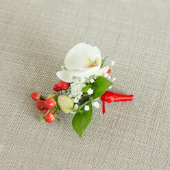 A single, elegant boutonniere is photographed from above on a light beige, woven fabric that looks like natural linen, its subtle texture adding a gentle, homely warmth reminiscent of reception venues around Gerrards Cross and the Chiltern countryside. Dominating the arrangement is a flawless white Phalaenopsis orchid, its petals broad and smooth like porcelain wings, spreading open with serene grace. At the heart of the bloom, a delicate wash of creamy yellow and muted orange glows softly, contrasting just enough to draw attention without overpowering the rest of the design. Encircling the orchid, wisps of baby's breath form a lace-like crown, each tiny white flower giving the piece an ethereal softness and creating a sense of lightness and air. A mix of lush, glossy green leaves, some long and tapering, others shorter and more elliptical, are carefully tucked around the blooms, offering structure and a fresh, garden-like feel. To one side, a tight cluster of bright, cherry-red berries adds a joyful splash of colour, their round, polished forms suggesting winter celebrations, Christmas services, or elegant weddings near West Common. A few paler green, bead-like buds sit alongside, suggesting growth and renewal. The stems are expertly wrapped in a vivid red satin ribbon, wound snugly and finished with a neat knot, the short tails resting with crisp precision. The overall effect is refined yet welcoming-a small floral accent that feels ideal for a groom or groomsman, a winter prom, or any formal occasion in the Gerrards Cross area where a touch of natural beauty and careful craftsmanship is cherished, and where local florists understand how a single flower at the heart can carry deep emotion.