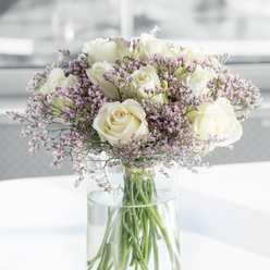 In this image, a refined sympathy bouquet of white roses and pale purple filler flowers stands gracefully in a clear glass vase, evoking the quiet streets and green calm of Gerrards Cross. The roses are the heart of the design, each bloom large and open, with layered, creamy-white petals that curl softly at the edges into classic spiral centers. Their colour is a gentle off-white, touched with hints of ivory, creating a mood of purity and dignified remembrance. Nestled around and between them are airy sprays of tiny lavender and lilac-pink blossoms, reminiscent of statice, giving the arrangement a light, cloud-like aura. These smaller flowers bring both texture and a subdued wash of pastel colour, mirroring the muted tonality of a soft, overcast day on West Common. The stems, neatly gathered and trimmed, are clearly visible through the tall cylindrical vase, reaching into clear water that reflects a glimmer of natural light. This transparency highlights the freshness of the flowers and the care with which they have been prepared. The vase rests on a smooth white surface that catches faint reflections of the blooms above, while the background is blurred into a luminous haze of whites and greys, suggesting a peaceful indoor setting. The bouquet feels like it might be placed in a quiet sitting room or chapel near the heart of Gerrards Cross, offering a gentle, respectful presence at a time of loss. The overall effect is one of soft comfort, understated grace, and a tender way to say, "We are thinking of you."