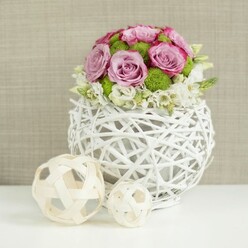 In this refined floral composition, a contemporary yet romantic centerpiece is captured as if set on a wedding table ready for guests arriving from across Gerrards Cross. A round white vase, created from slender, intertwined branches or rattan strips, forms a hollow globe with open gaps that let light filter through, giving the arrangement a delicate, almost weightless quality. Nestled tightly into this sculptural vessel is a domed bouquet of roses in soft blush pink and muted lavender, their layered petals curling gently outward, some tinged at the edges with richer, rosebud pink that hints at fragrance and warmth. Between these roses, clusters of vivid green button chrysanthemums emerge like tiny moss-covered hills, adding structure and a lively contrast that keeps the palette fresh rather than overly sweet. Around the base, fine white filler flowers-small, airy blossoms with slender stems-trail just slightly beyond the main outline of the bouquet, creating a soft, cloud-like fringe. Resting on the smooth white tabletop to the left are two coordinating decor pieces: openwork spheres woven from pale wicker or wood, one larger, one smaller, echoing the texture of the vase and reinforcing the handcrafted theme. Behind it all, a softly blurred, beige-toned fabric backdrop, reminiscent of natural linen or burlap, brings warmth without drawing attention from the flowers. The mood is serene and thoughtfully curated, ideal for a wedding breakfast at a local hotel near West Common or a stylish celebration at home, and it reflects the kind of balanced, textural design a local florist might create for couples marrying in and around Gerrards Cross.