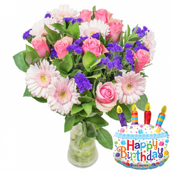 Sunlight slips around a joyful birthday bouquet, illuminating layers of pink and purple arranged with a florist's practiced hand - an ideal gift for a neighbour waiting on the Gerrards Cross station platform or a friend at the High Street café. The composition is dominated by fully opened pink roses, their velvety petals curling in delicate spirals that move from pale blush to a richer rose hue toward the heart of each bloom. Interspersed are generous pale-pink Gerbera daisies whose broad petals radiate outward, centres warmed by a soft yellow that feels almost buttery. Textural counterpoint comes from clusters of lively purple statice, each small, papery blossom lending a flicker of contrast and visual rhythm among the larger flowers. Deep green, glossy foliage frames the arrangement, creating a verdant backdrop that accentuates the fresh colours and suggests a cool, leafy scent when the bouquet is lifted. The group is arranged in a clear glass vase with a gently wider base; stems and water are plainly visible, reinforcing the impression of freshly-cut flowers prepared that morning. Nestled to the vase's lower right, a foil birthday-cake balloon complements the florals: white frosting with blue piped edging, scattered multicoloured sprinkles, and four tiny candles in purple, red, orange and blue with yellow flame tips. The words "Happy Birthday" appear in bright, mixed-colour lettering and a small KALEIDOSCOPE mark is visible at the balloon's foot. Together the blooms and balloon form a thoughtful, tactile surprise - a floral gift that reads as intimate celebration for someone in Gerrards Cross or nearby Beaconsfield.