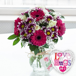Bathed in bright, natural light, this cheerful floral arrangement feels like it could be waiting on a kitchen table in Gerrards Cross, ready to greet someone coming in from the station. The flowers are gathered into a generous, rounded bouquet displayed in a sturdy clear glass vase, its transparency revealing carefully trimmed stems and crystal-clear water. Around the vase's neck, a piece of natural jute twine is tied in a relaxed bow, holding a small white tag decorated with a simple pink rose emblem and green handwritten-style lettering that hints at lovingly gathered flowers. The bouquet is led by several striking gerbera daisies in deep fuchsia or magenta, their wide petals framing rich, dark centres that draw the eye. Nestled between them are clusters of two-toned chrysanthemums, where velvety purple petals fade to bright white tips around golden-yellow centres, creating a lively, starburst effect. Creamy white roses, some still cupped in tight buds, add an air of classic elegance, while tiny white baby's breath blossoms scatter like soft lace through the arrangement, filling and softening the spaces. Fresh green leaves and foliage arc gently around the blooms, giving the design a full, lush profile like a garden border in full swing near West Common. To the right of the vase, slightly overlapping its base, sits a shiny heart-shaped foil balloon edged in silver, its white centre splashed with patterned letters spelling out "Love n' Hugs 24-7" in playful shades of pink, purple and blue. Set on a pale, uncluttered surface with a softly blurred, bright background, the image captures the warmth of a surprise flower delivery in Gerrards Cross, ideal for sending love, comfort, or everyday celebration.