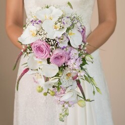 Viewed up-close and at eye-level, this cascading bridal bouquet captures the very spirit of romance as felt in Gerrards Cross's sun-dappled greens and elegant wedding venues. The bride, poised in a classic ivory gown with intricate lace, gently cradles her bouquet as if holding her own heartbeat-a testament to the anticipation and beauty of the day. The arrangement is a masterwork of texture and colour: pristine white Phalaenopsis orchids serve as the luminous anchors, their soft petals spilling gracefully and recalling the quiet blossoms under the chestnut trees at West Common. Intermingled are lush pink and lavender roses-some blooming wide, others curled in a tight embrace-each one a poetic echo of the gentle Surrey Lane gardens nearby. Wisps of baby's breath wend between the blooms, creating pockets of lightness whilst feathery Amaranthus in vibrant magenta weaves drama into the shape of the bouquet. The trailing effect is accentuated by subtle green stems and velvet-touch buds, a delicate nod to nature's unfolding in early summer. Interspersed throughout, tiny violets and lavender stock hint at a deeper depth, inspiring thoughts of shared vows and lifelong commitments. This handcrafted bouquet not only stands as a visual centrepiece but also captures the ambiance of Gerrards Cross-a place where fresh florals, local artistry, and love's quiet grandeur naturally intertwine.