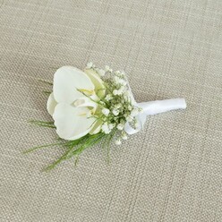 A refined white orchid boutonniere is laid out on a pale beige, woven fabric backdrop, its subtle threads and texture resembling an elegant linen table runner you might see at a Gerrards Cross reception. At the centre of the composition, a single Phalaenopsis orchid bloom opens serenely, its velvety white petals unfolding like smooth porcelain. Hints of soft yellow and cream nestle in the throat of the flower, drawing the eye inward and bringing a gentle warmth to the otherwise crisp palette. Surrounding the orchid, countless tiny blossoms of baby's breath create a misty, cloud-like collar, their delicate Gypsophila petals lending a sense of lightness and air. Threaded between these miniature flowers are wisps of vibrant green foliage, fine and feathery, giving the arrangement a fresh, garden-picked charm as though it were assembled from a hidden courtyard near West Common. The stems of the orchid, baby's breath, and greenery are carefully aligned and tightly bound with a smooth white satin ribbon, its neat wrapping and small, elegant bow suggesting the practiced hands of an experienced florist. The ribbon's gentle sheen catches the light, echoing the soft gloss of the orchid petals and tying the whole piece together visually and physically. The overall impression is one of understated sophistication and calm joy, perfect for a groom, best man, or proud father of the bride preparing for a ceremony in or around Gerrards Cross. This image captures not just a floral accessory, but the quiet ritual of getting ready, the boutonniere waiting patiently to be pinned to a lapel before the day's celebrations unfold.