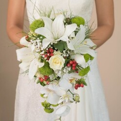 As you picture this radiant bridal moment, imagine a bride in a soft, flowing gown standing near the leafy avenues of Gerrards Cross, clutching an elaborate cascading bouquet that dazzles with artistry and detail. The bouquet's grandeur stems from the interplay of snowy white lilies, their star-shaped petals adorned with delicate speckles that suggest movement and vitality. Central to the ensemble is a creamy rose, its swirling petals tightly held-a symbol of everlasting romance illuminated by natural daylight filtering through the window. Trailing downward, a string of white phalaenopsis orchids lends the bouquet its signature teardrop silhouette, each bloom cascading effortlessly to evoke gentle motion. Here and there, vibrant green mums and textured foliage evoke the lush landscapes of Chalfont Park, adding freshness and modern vibrancy. Baby's breath, with its light airy clusters, intersperses softness and introduces a cloudlike frame around the larger blooms. Scattered within are festive clusters of bright red berries, reminiscent of the seasonal displays seen at Gerrards Cross markets, their cheerful presence providing lively contrast and joyous undertones. The entire bouquet is cradled in the bride's hands, a subtle ribbon visible at the handle. Every petal, leaf, and berry is revealed in soft natural light, capturing a spectrum of textures-from the satin sheen of the lilies to the velvet of the rose. Every detail speaks to the promise and hope woven into this singular moment at a Gerrards Cross wedding.
