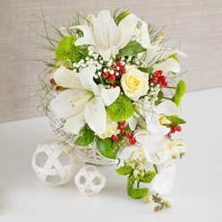 On a pristine white tabletop, an opulent floral arrangement in a round, white wicker basket radiates calm sophistication, as if prepared for an intimate wedding breakfast in Gerrards Cross. The eye is first drawn to the large white lilies that reign over the design, their petals wide open in full bloom, delicately speckled and framing golden-tipped stamens. Just above the centre, a tightly layered cream-to-pale-yellow rose provides a gentle glow, its spiralling petals suggesting quiet romance and care. From the right-hand side, a stream of white Phalaenopsis orchids cascades downward in a soft arc, each flat, elegant bloom adding to the bouquet's waterfall silhouette. All around, mixed greenery creates depth: fine, wispy blades, fern-like textures, and more substantial glossy leaves, intertwined with bright green pom-pom blooms that look almost like miniature tufts of moss. Throughout the arrangement, tiny white baby's breath flowers soften the lines, while bright red berries cluster between petals, their glossy skins offering a lively contrast, reminiscent of berries found along the leafy roads leading towards Chalfont St Peter. Two decorative white wicker orbs sit to the left on the table, repeating the basket's texture and lending a sculptural, modern feel to the composition. The background is a soft, textured beige fabric, gently lit so that no harsh shadows intrude, allowing the whites and greens to sing. The overall effect is one of purity, freshness, and celebration, a centrepiece that would feel perfectly at home gracing a wedding table or special family gathering in Gerrards Cross, crafted with the practiced eye of a local florist who understands both elegance and understated charm.