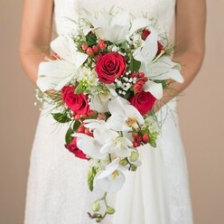 A radiant bridal bouquet steals the scene in this dreamy moment captured near Gerrards Cross. The bride's upper body glows against a softly blurred, warm beige backdrop, her arms and the delicate detailing of her lace wedding gown visible but secondary to the voluminous bouquet she holds. White lilies dominate the centre, their broad petals pervading the air with a subtle, familiar fragrance, while vivid crimson roses pulse with emotion, each blossom a symbol of devotion. Flowing downward are elegant white orchids-phalaenopsis, their graceful arcs and yellow-throated centers adding a sense of movement, as if echoing the gentle riverbanks that wind around East Common. Festive red berries glisten amidst the ivory and green, paired with frothy clusters of baby's breath that lighten the overall effect. Wispy ferns and emerald leaves form a lush, textured base, with a few spiky stems breaking the pattern, suggesting a blend of cultivated beauty and nature's spontaneity. The bouquet's dramatic cascade shape flows like the softest train of a wedding dress, creating a sense of anticipation and promise. Perfect for local weddings, this arrangement speaks softly of joy and tradition, echoing the community spirit shared by families in Gerrards Cross and nearby Chalfont St Peter.
