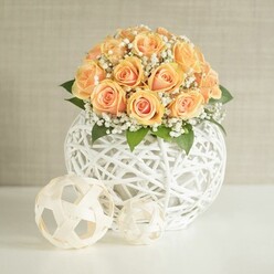An elegant floral centerpiece is captured in calm, even light, showcasing a domed bouquet of peach-toned roses that looks as though it has just been arranged by a careful hand. Around twenty to thirty roses, in shades ranging from soft apricot to warm honeyed orange, are nestled tightly together so that their curving petals overlap like a sea of silk. Some blooms are more open, revealing their deeper centres, while others are still cupped and shy, giving the arrangement a sense of natural progression and freshness. Between the roses, fine sprays of white gypsophila drift like mist, softening the edges and adding a gentle, cloud-like halo. Subtle touches of dark green foliage emerge from beneath the blooms, emphasising the rounded silhouette and giving the eye a moment of cool contrast. The flowers are cradled by a spherical white vase made from slender woven strips that criss-cross to form a light, airy lattice, allowing glimpses through to the interior and lending a sculptural, contemporary feel. On the pristine white surface in front of the vase, two smaller woven spheres in pale, natural tones sit playfully, repeating the lattice motif and tying the whole composition together. The background is a simple, softly textured wall in a pale beige or greige, typical of a calm, modern living space in Gerrards Cross, where natural light and neutral tones are prized. The overall effect is of quiet sophistication and gentle joy, perfectly suited to an intimate wedding breakfast at a local venue near West Common, an anniversary surprise delivered to a home off Oxford Road, or a refined gift arrangement prepared with local expertise by Flower Delivery Gerrards Cross for same-day delivery across the area.